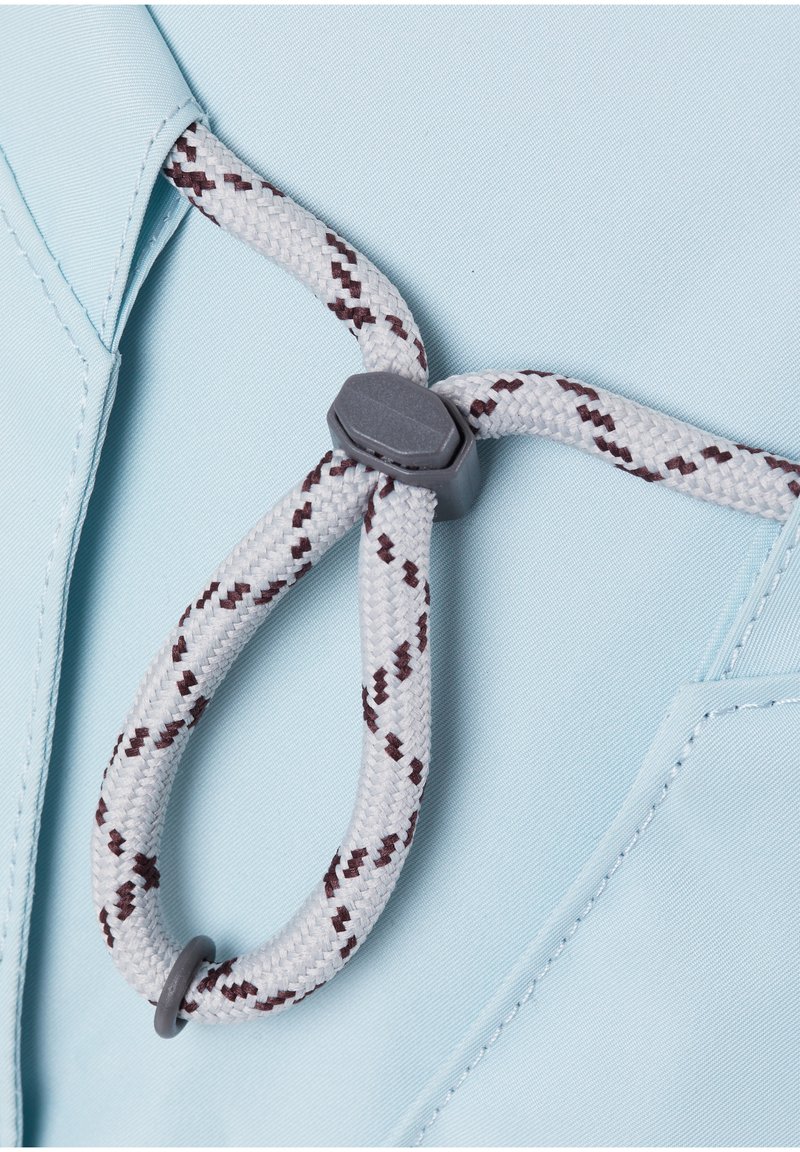 Primer plano de un cordón trenzado blanco y marrón con un tope de plástico gris sobre una tela azul clara con costuras visibles.