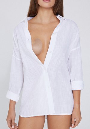 Camicetta bianca in lino con maniche lunghe, colletto aperto e chiusura con bottoni. Presenta un tessuto testurizzato e una vista parziale della pelle sottostante.