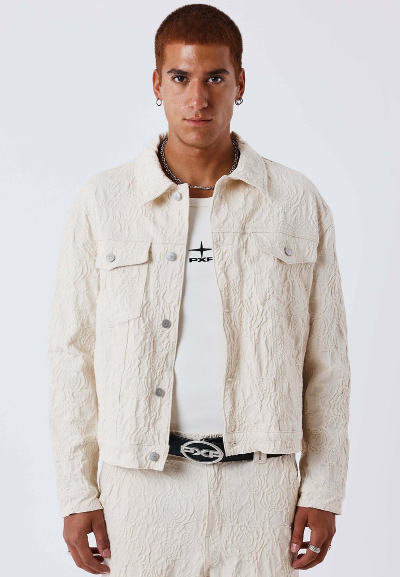 Jeune homme aux cheveux courts portant une veste et un pantalon texturés écru, une chemise blanche avec un logo noir, un collier chaîne, des boucles d'oreilles et une ceinture noire.