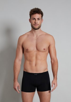 Schwarze Baumwoll-Boxershorts mit einer engen Passform, ausgestattet mit einem Markenbund. Das Modell ist oberkörperfrei und zeigt das Produkt deutlich.