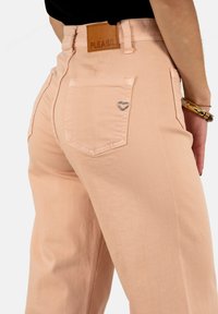 Los pantalones de mezclilla rosa claro cuentan con un bolsillo trasero adornado con un embellishment metálico en forma de corazón y una etiqueta de cuero marrón con la palabra "PLEASE" grabada.