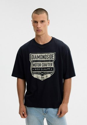 Un hombre luce una camiseta negra con un gran diseño de «Diamondside Motor Crafter», combinada con unos vaqueros azul claro, sobre un fondo blanco liso.