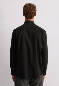 Man met kort krullend haar die een zwart shirt met lange mouwen en een grijze broek draagt, staande met het gezicht naar een plain witte muur.