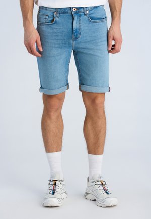 Mężczyzna noszący jasnoniebieskie, podwinięte szorty dżinsowe, białe wysokie skarpetki i białe trampki z wielokolorowymi akcentami.