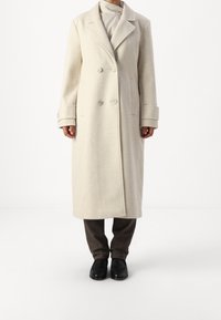 Cappotto in misto lana beige chiaro con design doppiopetto, rever a punta e grandi bottoni frontali. Presenta tasche laterali e polsini risvoltati.