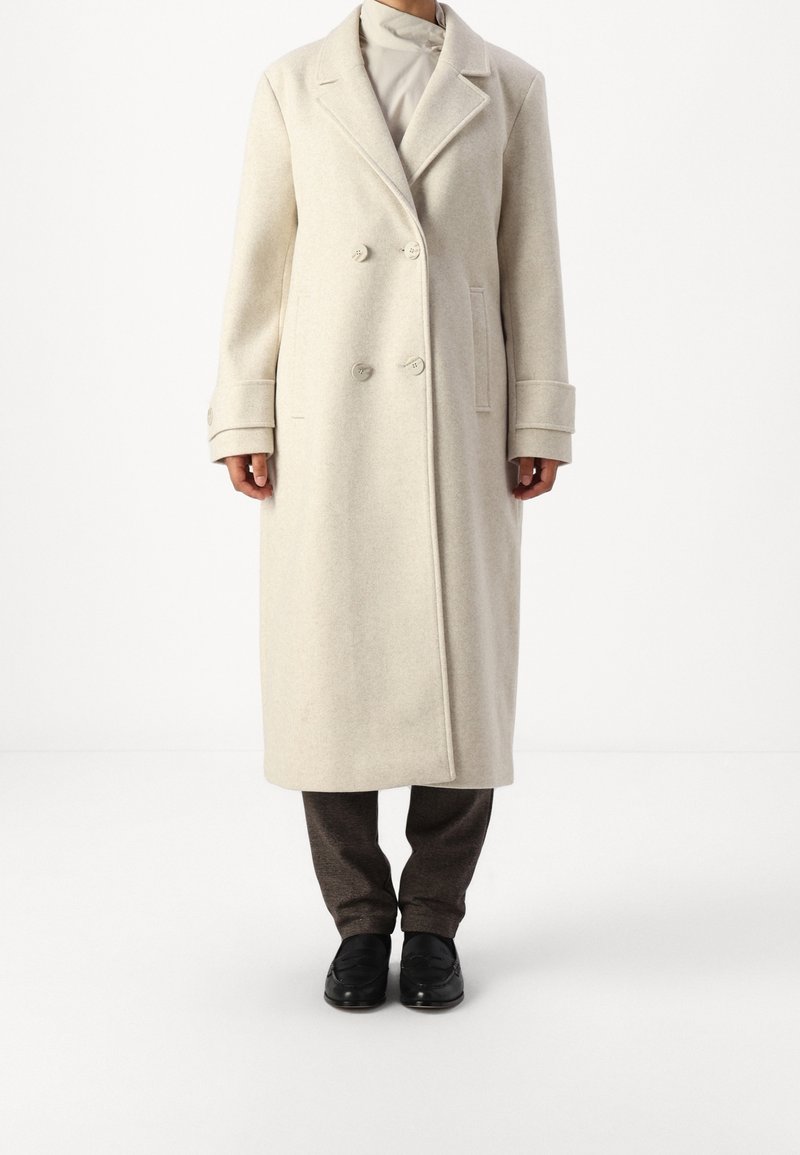 Cappotto in misto lana beige chiaro con design doppiopetto, rever a punta e grandi bottoni frontali. Presenta tasche laterali e polsini risvoltati.