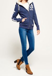 Felpa con cappuccio foderata in pile blu navy con grafiche bianche, chiusura con zip e cordini. Indossata con jeans skinny blu e stivali alla caviglia marroni.