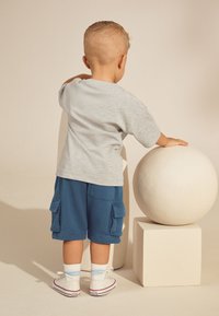 Gray cotton T-shirt, blue cargo shorts, white sneakers with red accents. Child stands beside a round sculpture on a pedestal.
