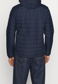 Marineblauwe gewatteerde jas met capuchon, met een gestructureerd oppervlak en elastische manchetten, gecombineerd met donkere spijkerbroeken.