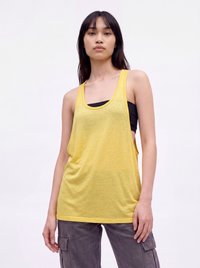 Femme aux longs cheveux foncés portant un haut jaune sans manches par-dessus un soutien-gorge de sport noir et un pantalon cargo gris, debout devant un fond blanc uni.