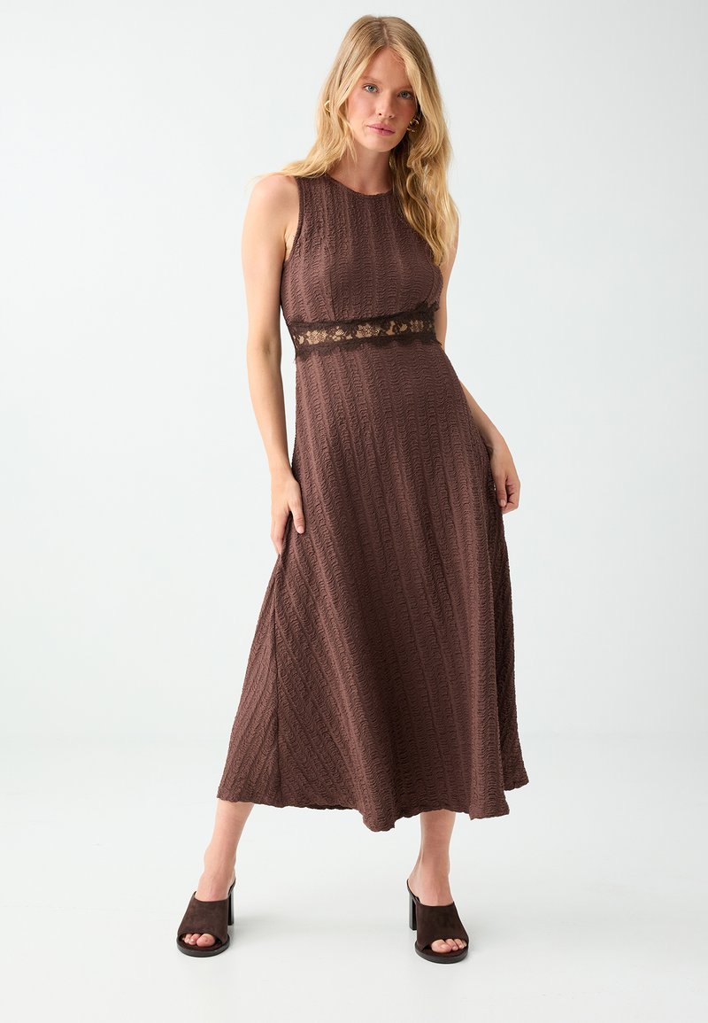 Femme portant une robe midi marron texturée sans manches avec un détail en dentelle à la taille et des sandales à talons ouvertes foncées, debout devant un fond uni.