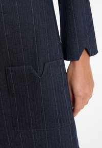 Nahaufnahme des Ärmels eines marineblauen Herringbone-Musters Mantels mit gestickter Tasche und einer Hand, die daneben ruht.