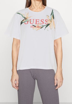 Femme portant un t-shirt blanc avec le texte "GUESS EST. 1981" et un motif floral sur les épaules, associé à un pantalon gris côtelé.