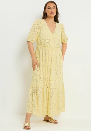 Kvinde stående iført en lysegul øjets maxi-kjole med pufærmer og V-hals, kombineret med guld- og beige flade sandaler.