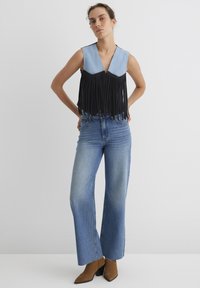 Schwarzes Fransenoberteil mit Denim-Details, V-Ausschnitt, Nietenakzenten, kombiniert mit hochtaillierten blauen Schlagjeans und braunen spitzen Ankle Boots.