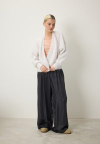 Cardigan blanc duveteux, haut rose clair et pantalon noir satiné ample avec une texture lisse. La mannequin se tient sur un sol en béton clair.