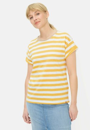 Kobieta o blond włosach, ubrana w żółto-białą koszulkę w paski z krótkim rękawem i jasnoniebieską spódnicę dżinsową na jednolitym tle.