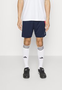 Marineblå sportshorts med en röd detalj, parat med vita knästrumpor med svarta ränder och logotyper, samt svarta fotbollsskor.