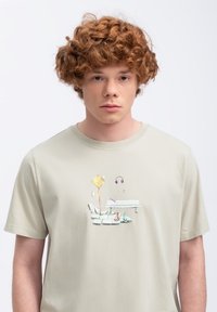 Lichtbeige katoenen T-shirt met een kleurrafische afbeelding van een bank, een bord, een hoofdtelefoon en schoenen. Korte mouwen en een ronde hals.