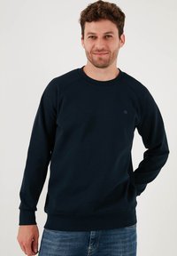 Námořnický modrý svetr vyrobený z měkké látky, s kulatým výstřihem, dlouhými raglánovými rukávy a malým logem na hrudi.