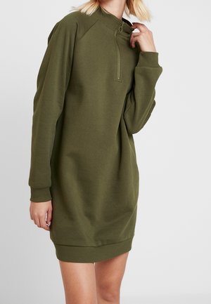 Kobieta w oliwkowej, długiej sukience typu sweatshirt z długim rękawem i kołnierzem z zamkiem do ćwiartki, stojąca na tle jasnego, jednolitego tła.