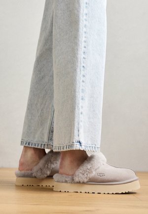 Pieds portant des chaussons UGG gris clair à enfiler avec une doublure duveteuse, associés à un jean denim bleu clair sur un sol en bois.