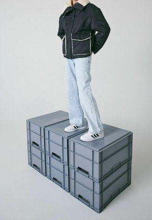 Schwarze isolierte Jacke mit cremefarbenem Kragen, zwei Fronttaschen; hellblaue Jeans; steht auf drei grauen Aufbewahrungsboxen. Weiße Sneakers mit schwarzen Streifen.