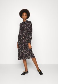 Vestido preto com flores, de mangas longas, com gola abotoada e cintura ajustada. Apresenta pequenos padrões de flores rosa e verde. Usado com mocassins pretos.