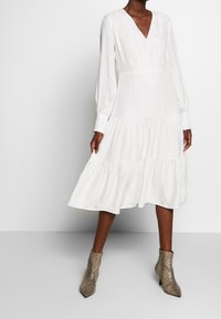 Biała sukienka z długim rękawem i dekoltem w serek, z koronkowymi akcentami na górze i warstwowym dołem. Połączona z teksturowanymi botkami w wężowy wzór sięgającymi kostki.