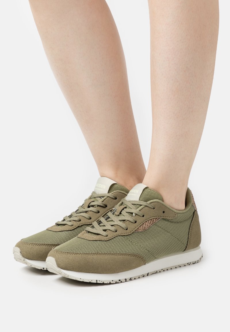 Paire de baskets légères en mesh et suède vert olive clair avec semelles blanches portées pieds nus, vues de côté et de face.