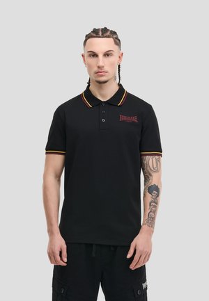Giovane con capelli intrecciati che indossa una polo nera Lonsdale con rifiniture rosse e gialle e pantaloncini cargo neri, mostrando tatuaggi sul braccio.