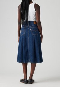 Falda midi de mezclilla en azul oscuro, con una cintura alta, pinzas, dos bolsillos traseros y una etiqueta de cuero marrón en la cinturilla. Zapatos planos negros.