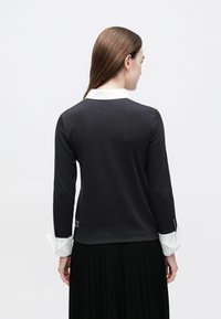 Žena s rovnými hnedými vlasmi má na sebe čierny top s dlhými rukávmi a bielym golierom a manžetami, doplnený čiernou plisovanou sukňou, otočená chrbtom.