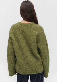 Olive zielony, oversize sweter o puszystej teksturze, okrągłym dekolcie i opadających ramionach, zestawiony z ciemnoniebieskimi dżinsami.