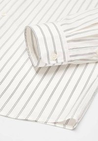 Chemise blanche à manches longues avec de fines rayures verticales noires, poignet plié montrant un bouton blanc, et un ourlet légèrement incurvé.