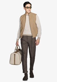 Beige lynlåsvest over en lys sweater, brune strukturerede bukser og mørke støvler; holder en creme farvet weekendtaske med brune detaljer.
