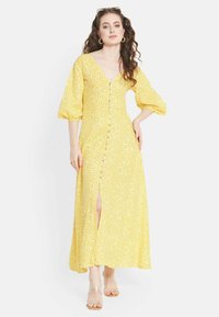 Gelbes, blumiges Maxikleid mit Puffärmeln, Knopfleiste und Schlitzen bis zum Oberschenkel. Hergestellt aus leichtem, strukturiertem Stoff. Klare Sandalen.