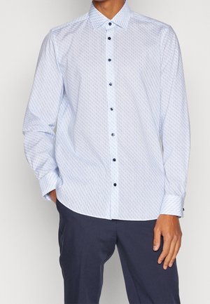 Man wearing light blue patterned long-sleeve button-up shirt with dark buttons and dark navy pants, standing with one hand in pocket.