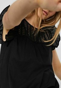 Giovane donna che indossa una blusa nera con dettagli in pizzo e maniche a balze, che si protende in avanti con il braccio teso.