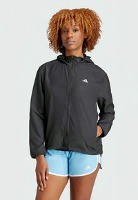 Eine Frau mit lockigem Haar steht mit einer Hand in der Tasche, trägt eine schwarze, mit Reißverschluss versehenen Kapuzenjacke und hellblaue Sportshorts.