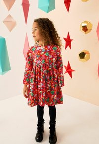 Rode bloemenjurk met lange mouwen, voorzien van een geraffineerde taille. Gecombineerd met zwarte leggings en glanzende zwarte laarzen. Kleurrijke papieren versieringen op de achtergrond.