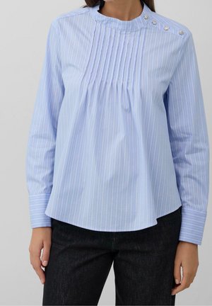 Mujer vistiendo una blusa azul claro con rayas finas, con pliegues en la parte delantera y botones decorativos en el hombro, combinada con pantalones oscuros.