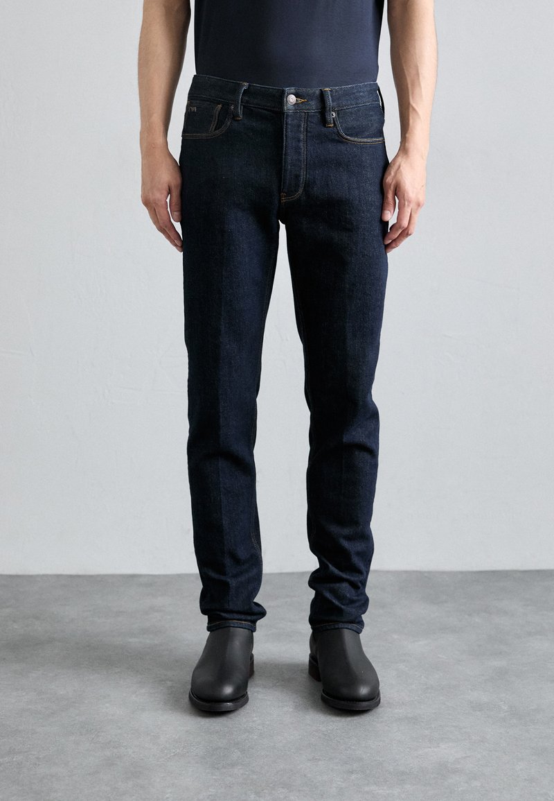 Jeans ajustados de color azul oscuro con un diseño clásico de cinco bolsillos, sutiles detalles de costura, combinados con botines negros en un suelo gris.