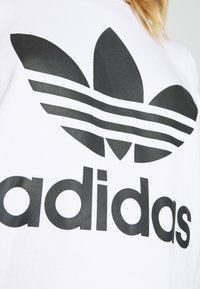 Bela dukserica s velikim crnim Adidas logotipom s tri lista poput pruga i podebljanim tekstom ispod. Izrađena od glatke pamučne tkanine.