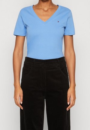 Woman wearing a light blue V-neck short-sleeve shirt and high-waisted black corduroy pants, standing with arms relaxed.