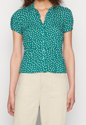 Kobieta ubrana w zieloną bluzkę z krótkim rękawem, białym kwiatowym nadrukiem i wyciętą falbanką przy kołnierzyku, zestawioną z jasnobeżowymi spodniami.