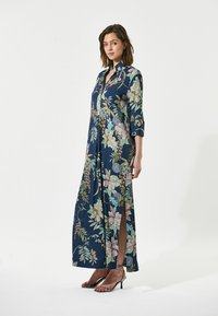 Abito maxi in tessuto blu navy con motivi floreali e fogliari, colletto alto e spacchi laterali. Modella che indossa sandali con tacchi alti argento.