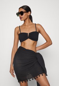 Črn bikini zgornji del z teksturirano tkanino, ki ima podrobnost vozla spredaj. Prosojna črna sarong s čipkastimi detajli se ovija okoli pasu.
