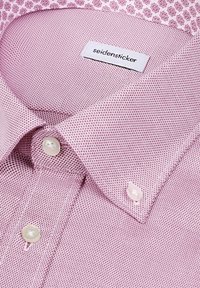 Gros plan sur une chemise plissée rose à motifs avec des boutons blancs et une étiquette de marque "seidensticker" visible à l'intérieur du col.
