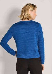 Frau mit kurzem blonden Haar, die einen strukturierten blauen Langarmpulli und schwarze Hose trägt, von hinten vor einem schlichten Hintergrund betrachtet.